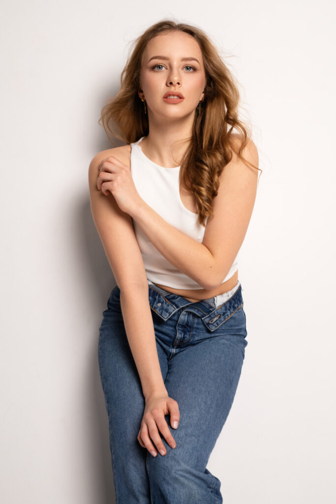 Młoda kobieta w białym topie i dżinsach, z długimi, falującymi włosami, pozująca w neutralnym tle. Patrzy świadomie w obiektyw, lekko przytrzymując ramię