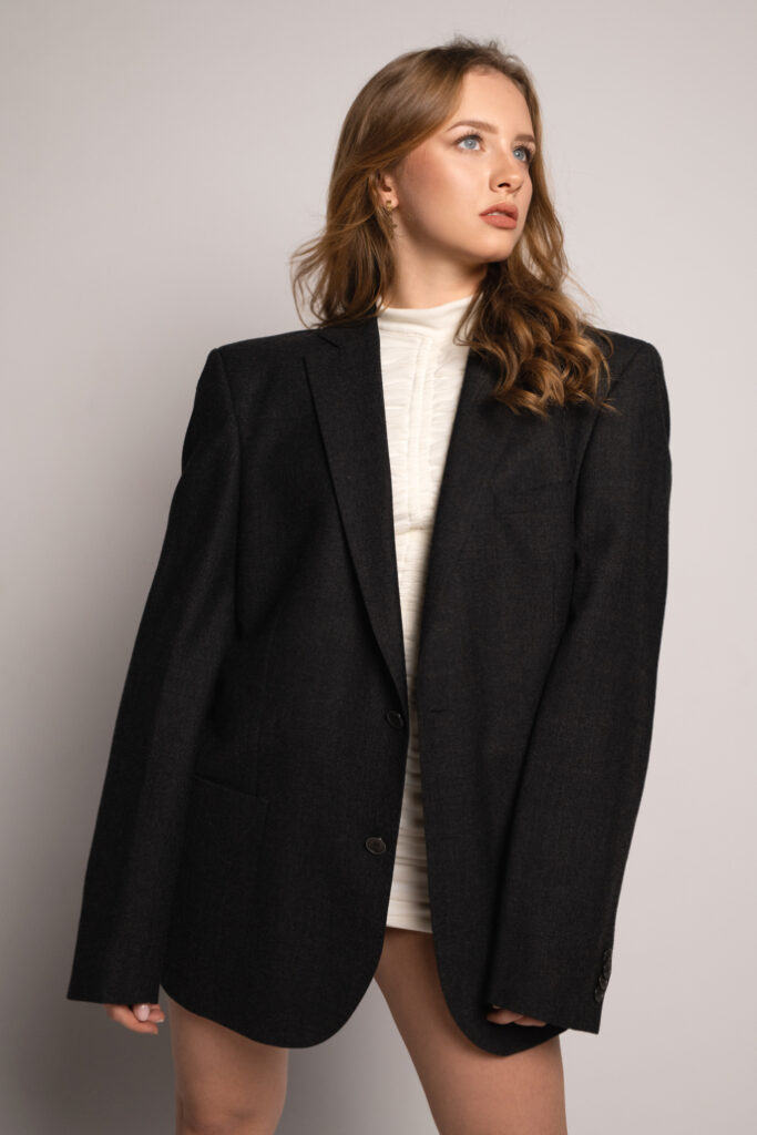 Kobieta w oversize'owym czarnym żakiecie zestawionym z jasną, dopasowaną sukienką, pozuje z pewnością na jasnym tle