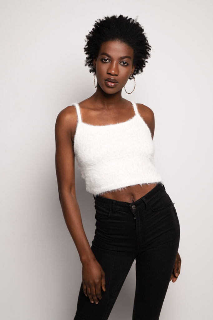Młoda kobieta o krótkich, kręconych włosach, ubrana w biały, futrzany top i czarne, dopasowane spodnie. Stoi na jasnym tle, patrząc bezpośrednio w obiektyw, z jedną ręką na biodrze