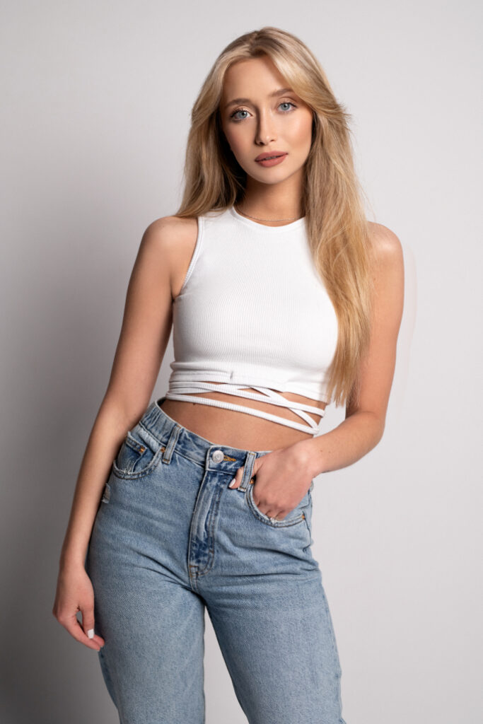Młoda kobieta w białym topie i niebieskich dżinsach stoi na jasnym tle, lekko odwracając się w bok i uśmiechając