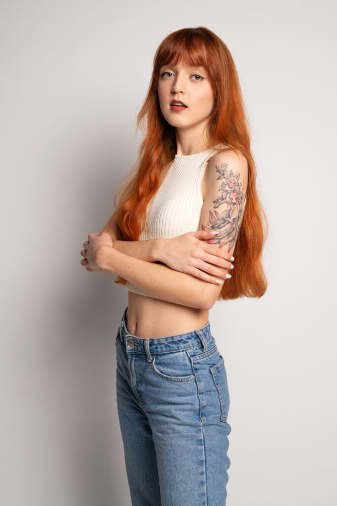 Młoda kobieta z długimi rudymi włosami, w białej bluzce bez rękawów i niebieskich dżinsach, pozująca na neutralnym tle. Na jej ramieniu widoczny jest kolorowy tatuaż