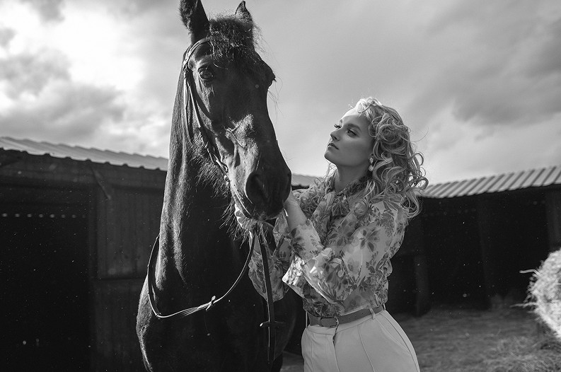 Kobieta z kręconymi włosami w białej bluzce stoji obok czarnego konia, z delikatnie uniesioną głową, w tle widać drewniane budynki i chmury