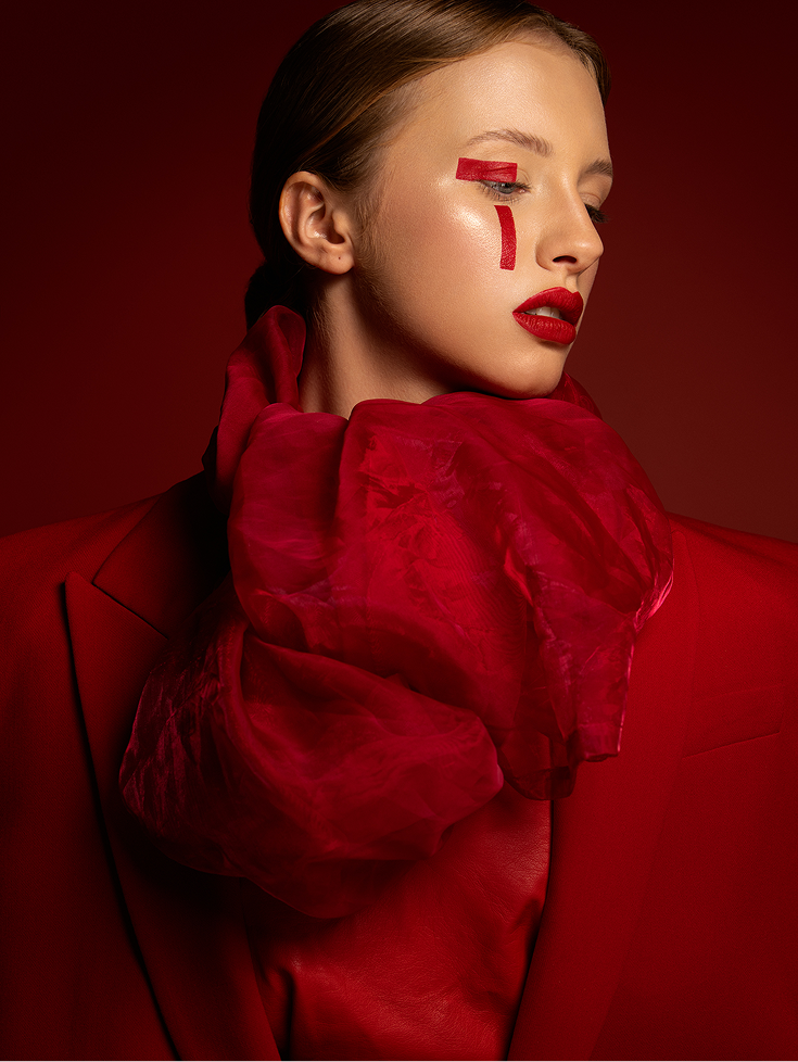Kobieta w intensywnie czerwonym ubraniu, z blousem i dużym szalem. Makijaż z czerwonym akcentem na policzku i ustach. Tło w odcieniu czerwonym