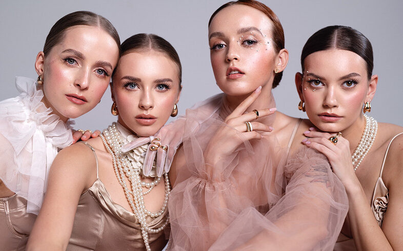 Cztery kobiety pozujące razem, każda w eleganckich strojach w odcieniach beżu i różu, z wyrafinowanym makijażem. Dwie z nich mają na sobie perły, a wszystkie noszą biżuterię. W tle neutralny kolor