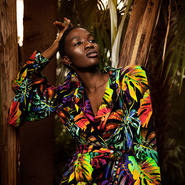 Kobieta w kolorowej, egzotycznej sukience z motywem roślinnym, stojąca w tropikalnym otoczeniu z palmami. Ma delikatny wyraz twarzy i jest w lekko zrelaksowanej pozie