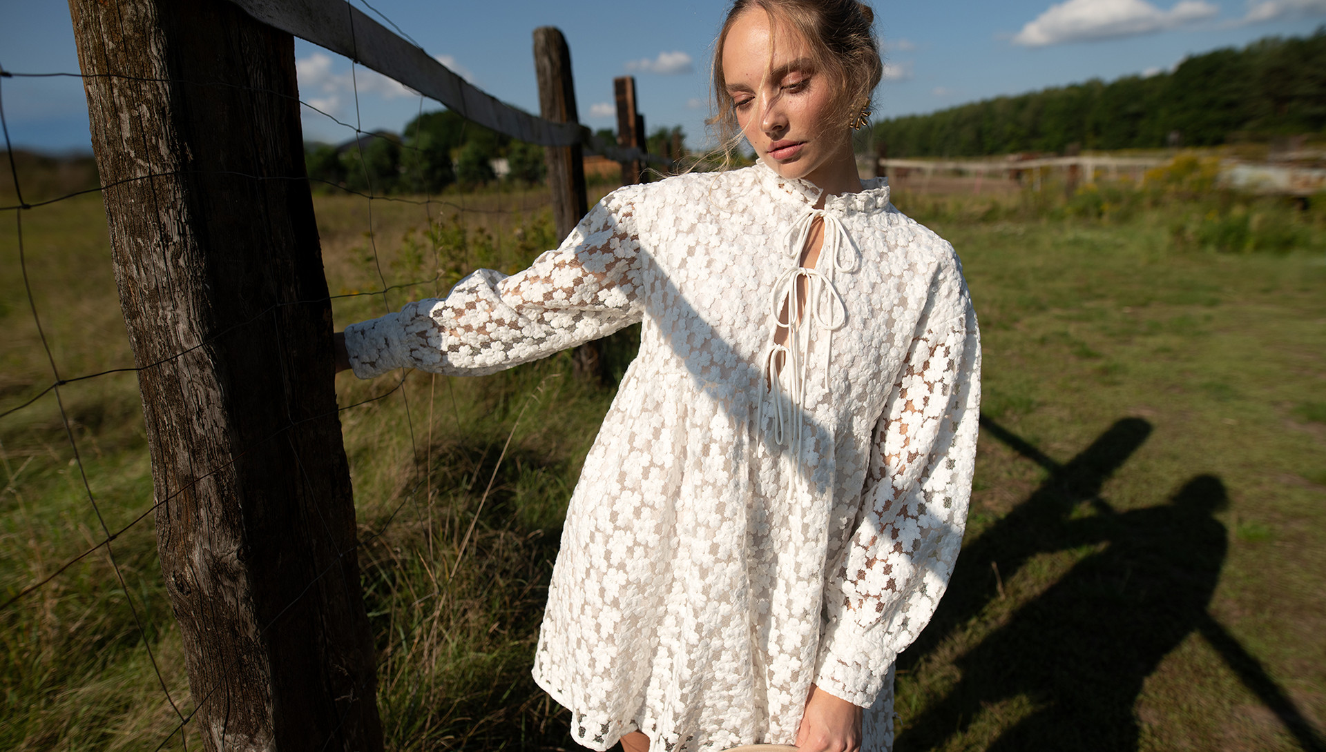 Kobieta w białej, koronkowej sukience stoi obok drewnianego płotu na zielonej łące. Na twarzy ma delikatny wyraz. W tle widać drzewa i niebo z białymi chmurami